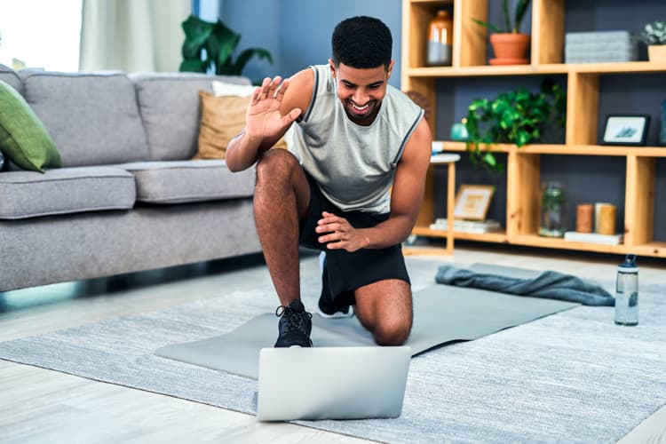 man in workout clothing kneeling on yoga mat and waving at laptop computer on floor